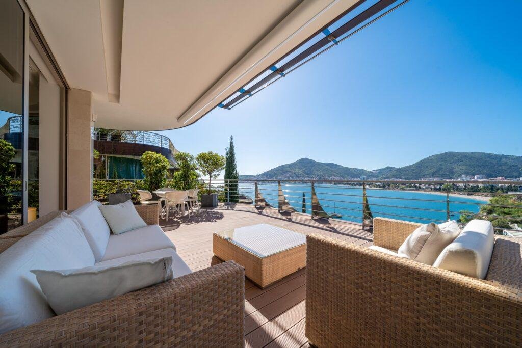 The terrace of a first-line apartment in the complex, featuring spacious and comfortable outdoor furniture, greenery on the terrace, and a view of the sea and Budva in the distance.