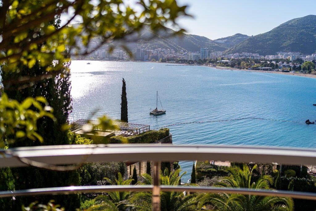 View from the terrace showing part of the railing, the sea, a sailboat, and Budva in the distance.