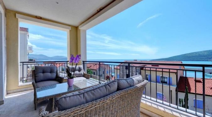 pogled na more i zgrade koje su u redu blizem moru u marina village, lustica bay. terasa ima pleteni namestaj i sive jastuke