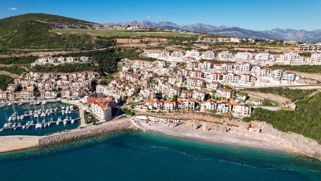 drone footage of close up marina village in lustica bay. densly built buildings, beach in front of them and also marina with yachts . in the back there a hill with untouched nature that surrounds marina village.