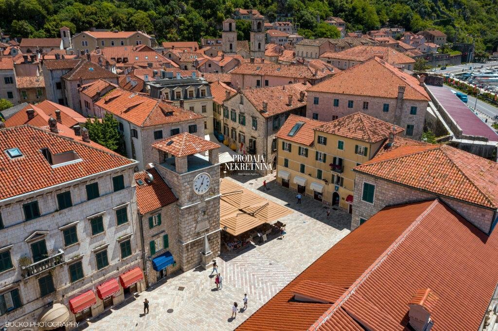 An aerial view shows Kotor Old Town on a beautiful sunny day, featuring the clock tower, numerous red-tiled rooftops, and stone façades. Several people can be seen walking through the town.