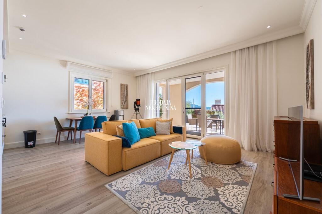 living room with bright yellow furiture and blue pillows. there is part of dining room. small table and four chairs are next to side window and there is also floor to ceiling windows for exiting to the terrace.