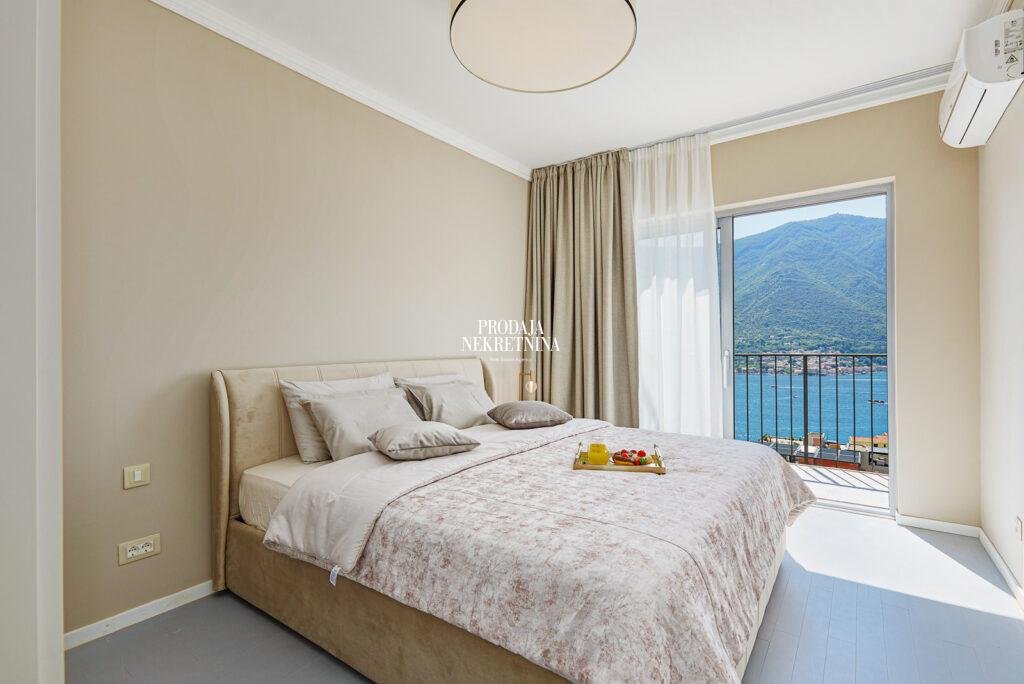 spavaća soba sa krem krevetom koji je centralno u kadru, a iza se nalazi prozor od poda do plafona sa svetlim zavesama i pogledom na more i brda