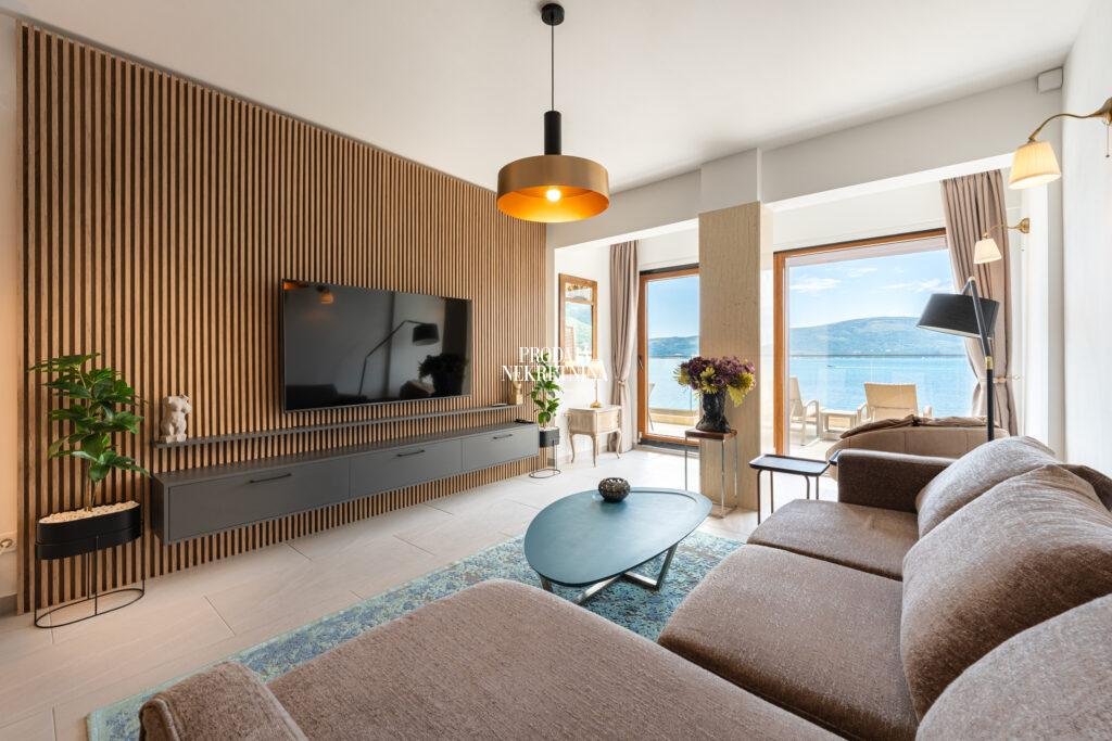 dnevna soba luksuznog staba sa vekikom braon garniturom, plavim tepihom, drvenim panelom preko celog zida na kojem je nakačen tv. dnevna soba ima stakla od poda do plafona čitavom širinom i odatle se vidi terasa i potpuno otvoren pogled na more