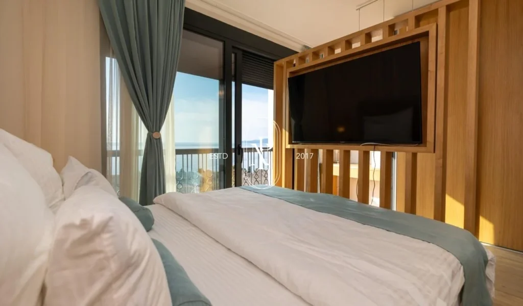 spavaća soba vile koja ima hotelski stil. Krevet ispred koga je drveni zidni panel na kojem se nalazi tv. iz kreveta se gleda direktno na prozor od poda do plafona sa pogledom na more