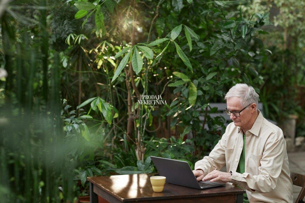 an older man sitting at the table with his computer, surrounder with lush vegetation. he seems focused, working.